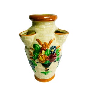 May include: A cream-colored ceramic wall pocket vase with a textured basket-weave pattern. The vase features a brown rim and base, with a central hand-painted floral design in shades of green, yellow, red, and blue, and two side openings.