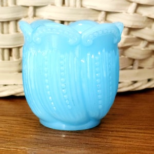 May include: A light blue, vintage-style glass vase. The vase has a tulip-like shape with a scalloped rim and vertical beaded accents. The vase is sitting on a wooden surface.