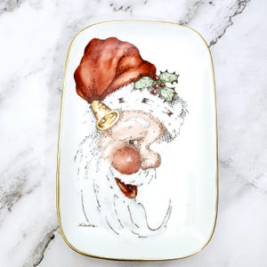 May include: Rectangular white ceramic dish with a gold rim, featuring a hand-painted Santa Claus design. Santa wears a red hat, has a white beard, and a gold bell. The dish is signed "Sandra."