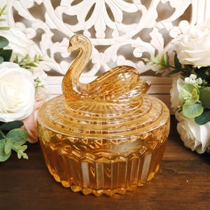 May include: A round amber-colored glass trinket dish featuring a swan-shaped lid handle. The base of the dish has a vertical ribbed pattern, while the lid displays a sunburst design radiating from the swan figure, crafted in translucent glass.