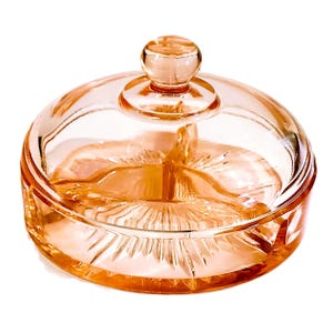 May include: A vintage, peach-colored glass candy dish with a lid. The dish has a round shape with a decorative, etched pattern on the base and a small knob on the lid. The glass is transparent, allowing light to pass through.