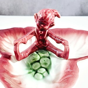 May include: A vintage ceramic serving dish with a whimsical design. The dish features a red elf figurine perched atop a cluster of green grapes. The dish itself has a petal-like shape with red and white coloring.