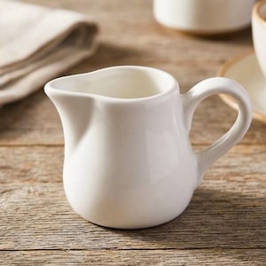 May include: A small, white ceramic creamer pitcher with a rounded body, a curved handle, and a distinct pouring spout. The pitcher features a smooth, glossy finish and a simple, functional design intended for serving cream or milk at the table.