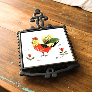 May include: Black metal trivet with a square white tile featuring a colorful rooster illustration. The rooster has red, yellow, and green feathers. Red tulip illustrations are also on the tile. The trivet has a decorative handle.