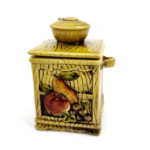 May include: A ceramic jar with a lid shaped like a small bowl. The jar is decorated with a woodgrain pattern and a painted scene of fruit, including an apple, pear, and grapes. The jar has a handle on the side.