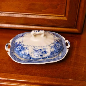 May include: A rectangular ceramic butter dish with a lid, featuring a white base and a blue floral pattern. The dish has two side handles and a central knob on the lid, with a blue cross-hatched border design around the edges of the base and lid.
