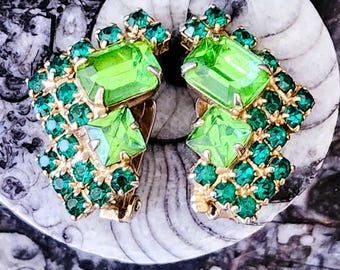 May include: A pair of vintage clip-on earrings. Each earring features a large, rectangular, light green gemstone surrounded by smaller, round, dark green gemstones. The earrings have a gold-tone metal setting and are displayed on a white and gray background.