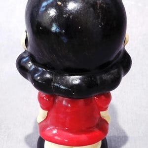 May include: A vintage figurine of a woman, viewed from the back. The figure has black hair and is wearing a red dress with a white collar. The base is black, and the figurine has a glossy finish.