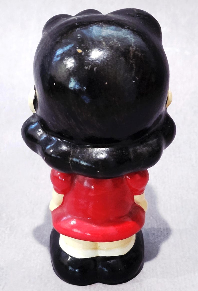 May include: A vintage figurine of a woman, viewed from the back. The figure has black hair and is wearing a red dress with a white collar. The base is black, and the figurine has a glossy finish.