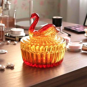 May include: A round glass trinket box with a lid featuring a sculpted swan handle. The glass transitions from a deep red base to a vibrant orange top, showcasing a textured, faceted pattern across the entire surface of the container.
