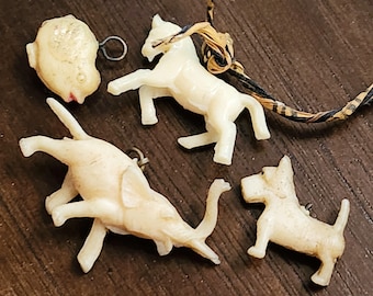 May include: A collection of small, off-white plastic animal charms, including a horse, elephant, dog, and a fish-shaped charm. Each charm has a small metal loop for attachment. The charms are arranged on a dark brown wooden surface.