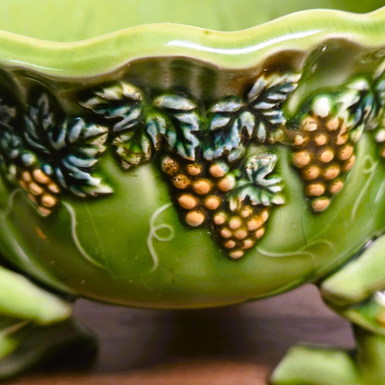 May include: A light green ceramic bowl featuring a raised relief pattern of grape clusters and vine leaves around the exterior. The bowl has a scalloped rim and stands on three decorative feet, with a glossy finish and visible fine crackle texture.