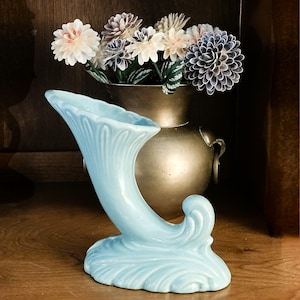 May include: A light blue ceramic cornucopia vase with a curved, horn-like shape. The vase has a decorative, leaf-like base and a fluted design on the opening. A brass vase with flowers is in the background.