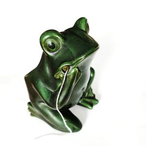 May include: A ceramic frog figurine in a glossy, dark green glaze. The frog is in a seated position with its hand near its face, and a white string is coming out of its mouth. The figurine has detailed features and is set against a white background.