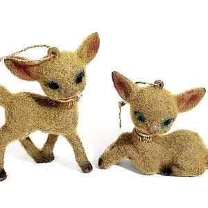 May include: Two vintage deer ornaments. One is standing, the other is lying down. Both have tan fur, pink ears, and blue eye shadow. They have gold-colored collars and hanging loops.