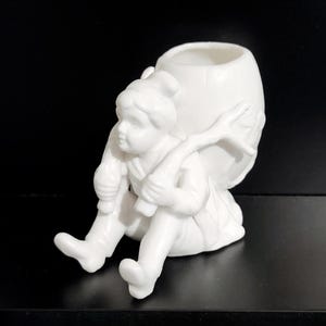 May include: A white ceramic figurine of a seated child carrying a large, open-topped vessel on their back. The child is depicted with detailed facial features and clothing, holding a branch. The figurine is a decorative item.