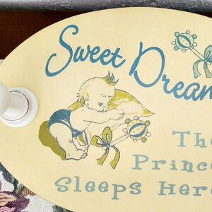 May include: Oval-shaped, light yellow sign with the text "Sweet Dreams" and "The Prince Sleeps Here" in teal. A sleeping baby illustration and rattle are also featured. Two white knobs are attached.