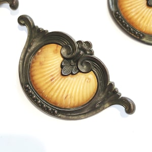 May include: Antique bronze-toned cabinet hardware with a seashell design. The hardware features a cream-colored, fan-shaped insert within an ornate, scrolling frame. The frame is detailed with small, round accents and floral embellishments.