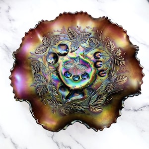 May include: An iridescent glass bowl with a ruffled edge. The bowl features a detailed embossed design of cherries, leaves, and other fruit. The colors shift from purple to gold and green, creating a shimmering effect.