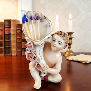 May include: A porcelain cherub figurine holding a floral-shaped vase filled with matches. The vase is white with gold trim and pink roses. The cherub is kneeling, with a serene expression. Two lit candles and a brass candlestick are in the background.