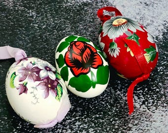 Pode incluir: Três ovos de Páscoa decorativos pintados à mão. Um ovo é branco com flores roxas, outro é branco com uma rosa vermelha e folhas verdes, e o terceiro é vermelho com um desenho floral branco e cinza. Cada ovo tem uma fita para pendurar.