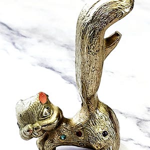 May include: A gold-colored squirrel-shaped bottle opener with a textured finish. The squirrel has a red gem on its head and colorful gems on its body. The tail curves upwards, and the opener is located in the tail.