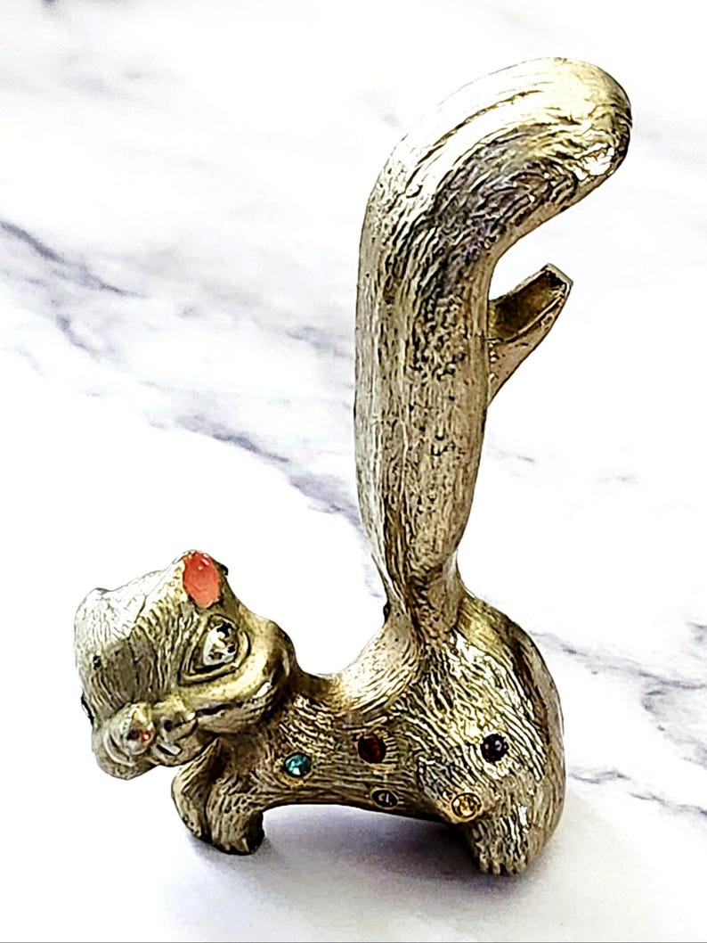 May include: A gold-colored squirrel-shaped bottle opener with a textured finish. The squirrel has a red gem on its head and colorful gems on its body. The tail curves upwards, and the opener is located in the tail.