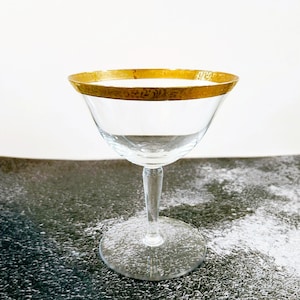 May include: A clear glass champagne coupe with a gold rim. The rim has a decorative pattern.