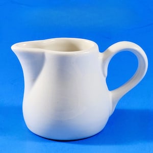 May include: A white ceramic creamer with a rounded body and a handle. The creamer is designed for pouring liquids.