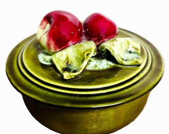 May include: A ceramic lidded dish in shades of green, adorned with a cluster of red cherries and green leaves. The cherries have a glossy finish, and the leaves have a textured appearance. The dish is round with a slightly raised base.
