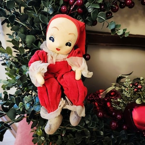 May include: A vintage doll with a painted face, wearing a red velvet outfit with white ruffled trim. The doll is part of a Christmas wreath with red berries and green foliage.