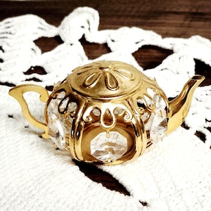 May include: A gold-colored teapot with a decorative filigree design and clear crystal accents. The teapot has a lid with a floral design and a curved handle and spout. The teapot is sitting on a white lace doily.