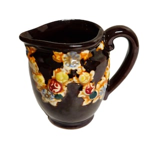 May include: A dark brown ceramic creamer pitcher featuring a raised, colorful floral pattern in shades of yellow, red, blue, and white. The pitcher has a curved handle, a wide pouring spout, and a smooth, glossy finish throughout the exterior.