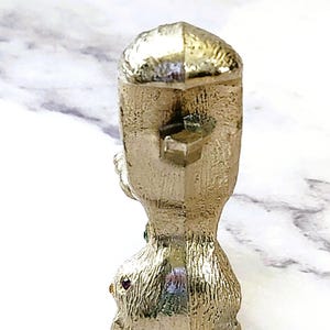May include: A gold-colored, textured figurine with a geometric design. The figurine has a square top and a rounded base. Small red and green gemstones are set into the base. The figurine is likely a decorative item.