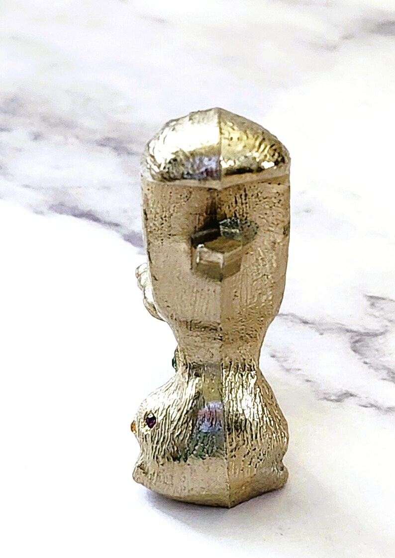 May include: A gold-colored, textured figurine with a geometric design. The figurine has a square top and a rounded base. Small red and green gemstones are set into the base. The figurine is likely a decorative item.
