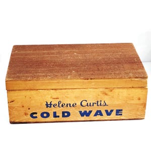 May include: A vintage wooden box with a hinged lid. The box is light brown with a darker brown lid. The words "Helene Curtis COLD WAVE" are printed in blue on the front of the box. The box appears to be in good condition.