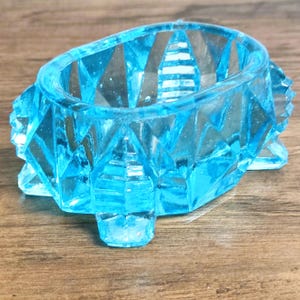 May include: A light blue, faceted glass dish with a geometric design. The dish has an oval shape with a textured rim and sits on three small feet. The glass is transparent, allowing light to pass through.