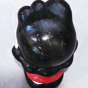 May include: A small, vintage figurine of a character with a black head and body, a red band around the middle, and white accents. The figurine is viewed from above, showcasing its glossy finish and simple design.