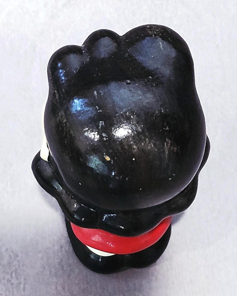 May include: A small, vintage figurine of a character with a black head and body, a red band around the middle, and white accents. The figurine is viewed from above, showcasing its glossy finish and simple design.