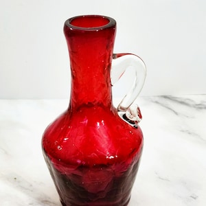 May include: A red glass pitcher with a clear glass handle. The pitcher has a rounded base and a narrow neck. The glass has a textured appearance. The pitcher is likely used for serving beverages.