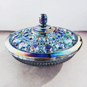 May include: Iridescent glass candy dish with a lid. The dish is round with a detailed geometric pattern and a silver rim. The lid has a small knob and a similar iridescent finish, reflecting blues, greens, and purples. The dish is likely vintage or antique.