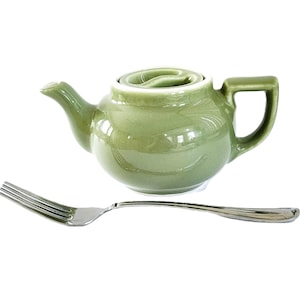May include: A small, sage green ceramic teapot with a matching lid and a square handle. A silver fork is positioned below the teapot. The teapot has a glossy finish and a curved spout. The background is white.