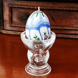 May include: An egg-shaped candle with a blue and white floral design, resting in a clear glass egg cup. The candle has a wick at the top and is decorated with green and pink accents. The egg cup is clear glass.