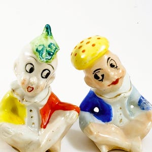 May include: A pair of vintage ceramic figurines, each depicting a whimsical character. One figure wears a yellow and red outfit with a green leaf-shaped hat. The other has a blue and yellow outfit with a yellow hat with red dots. Both have painted facial features.