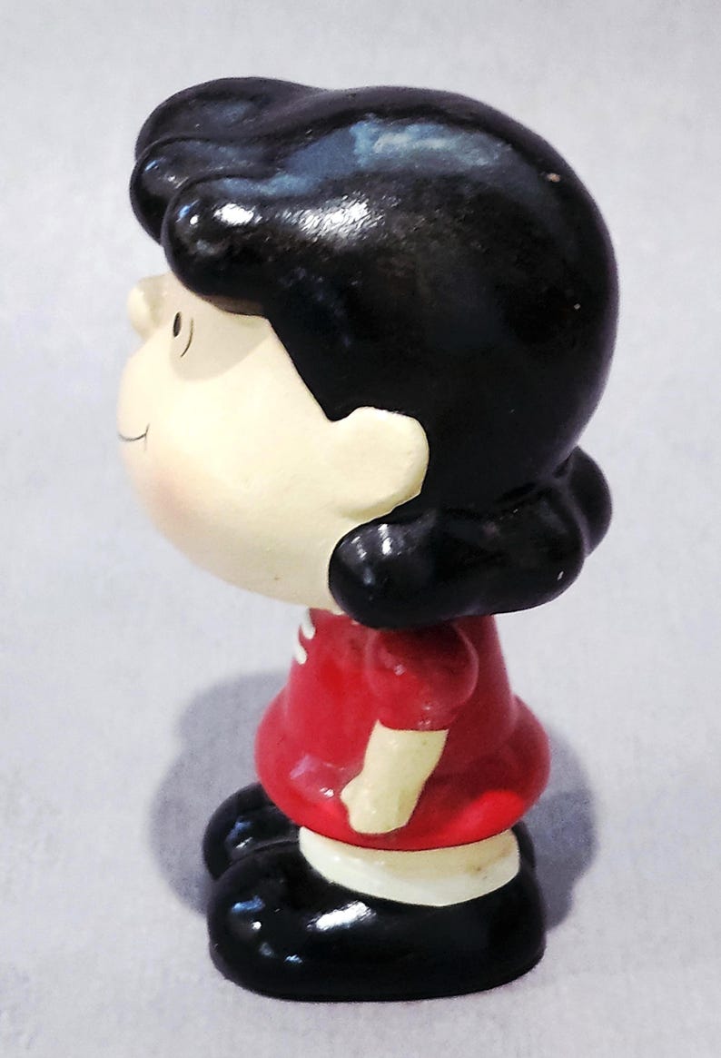 May include: A ceramic figurine of a cartoon character, featuring a black bob hairstyle and a red dress. The figurine has a white face with rosy cheeks and black shoes. The figure is a collectible item.