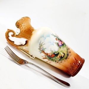 May include: A porcelain vase with a cream-colored body, a flared rim, and a gold-toned handle. The vase features a painted pastoral scene of figures in a landscape. A silver-toned metal fork is positioned alongside the vase for scale reference.