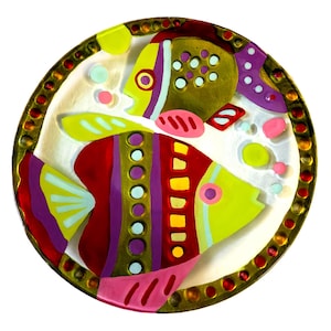 May include: A round decorative plate featuring two stylized fish in vibrant shades of red, yellow, purple, and pink. The border is gold with embedded red and bronze circular accents. The background is white with scattered colorful dots.
