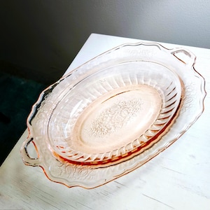 May include: A vintage pink glass serving dish set. The set includes an oval-shaped dish with a floral design and a larger, scalloped-edge dish. The dishes have handles and are made of transparent pink glass. The set is likely used for serving food.