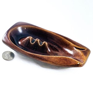 May include: A brown ceramic ashtray with a curved design. The ashtray has three cigarette rests and a smooth, glossy finish. A U.S. quarter is next to the ashtray for size comparison. The ashtray is a vintage item.