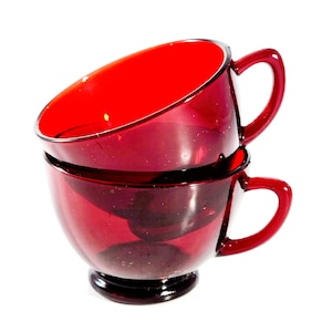 May include: Two stacked ruby red glass teacups with handles. The cups are slightly tilted and the top cup is partially obscured by the bottom cup.